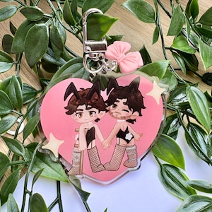 Peut inclure: Un porte-clés en forme de cœur avec deux personnages de dessins animés aux oreilles de lapin, se tenant la main et portant de la lingerie. Le cœur rose a des étoiles et est attaché à un fermoir argenté avec un nœud rose. Le fond est un mélange de feuilles vertes et de blanc.