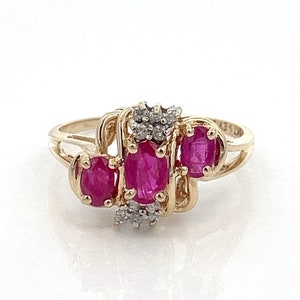 May include: A gold ring with three oval-shaped ruby gemstones and a cluster of small diamonds. The ring has a delicate, intricate design.