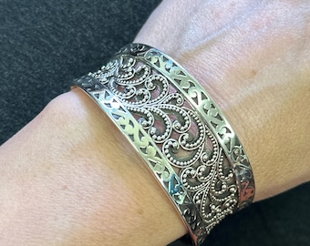 May include: A silver cuff bracelet with intricate, ornate scrollwork and dotted patterns. The wide band features a detailed design with openwork edges, creating a textured and elegant accessory. The bracelet is worn on a wrist.