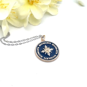 May include: A silver necklace with a circular pendant. The pendant is navy blue with a gold-colored border and features the word "HOGWARTS" in gold. The center has a gold emblem with clear crystals. The necklace is displayed on a white surface with a yellow flower.