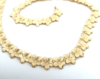 May include: A gold necklace with a chain made of small, detailed flower charms. The necklace is laid out in a loop on a white surface.