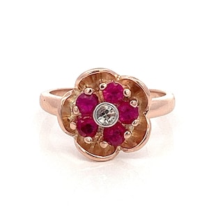 May include: A rose gold ring with a flower design. The flower has six red gemstones and a single white gemstone in the center.