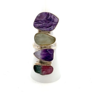 May include: A silver ring with four different colored stones. The stones are purple, green, pink, and white. The ring is on a white display stand.