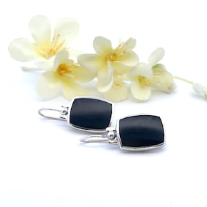 May include: A pair of silver earrings with a rectangular, black inlay. The earrings are set against a white background with soft yellow and white flowers. The earrings have a simple, elegant design.