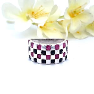 May include: A silver ring with a checkerboard pattern of red and black gemstones. The ring is wide and has a polished finish. The gemstones are square-cut and set closely together. The background features white flowers.