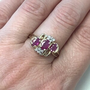 May include: A gold ring featuring three oval-shaped ruby gemstones and smaller diamond accents. The ring is set in a delicate, interwoven design.