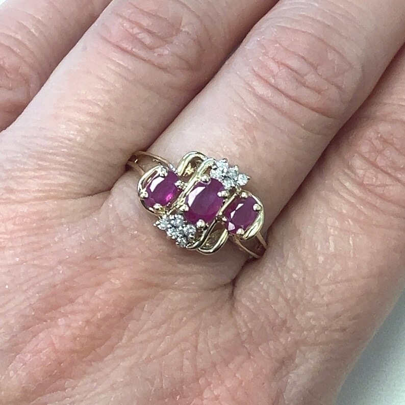 May include: A gold ring featuring three oval-shaped ruby gemstones and smaller diamond accents. The ring is set in a delicate, interwoven design.