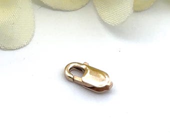 May include: A small, gold-colored lobster clasp, designed for jewelry making. The clasp is oval-shaped with a spring-loaded mechanism for secure closure. The metal has a polished finish, reflecting light. The background is white with a soft, blurred floral element.