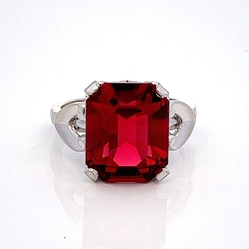 May include: A silver ring featuring a large, red, emerald-cut gemstone.