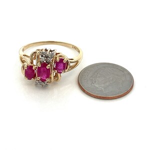 May include: A gold ring featuring three oval-shaped ruby stones and smaller diamond accents. The ring is shown next to a UK ten pence coin for size comparison.