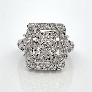 May include: A silver ring with a square-shaped setting featuring a large, round, clear center stone surrounded by smaller, clear stones.