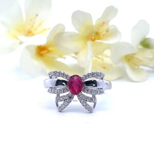May include: A silver bow-shaped ring with a central oval ruby and small clear stones. The ring is set against a backdrop of white flowers with yellow centers, creating a delicate and elegant aesthetic.