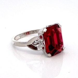May include: A silver ring featuring a large, red, emerald-cut gemstone. The ring has a heart-shaped design on the band.