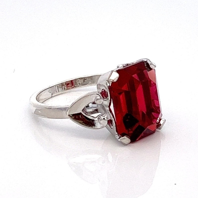 May include: A silver ring featuring a large, red, emerald-cut gemstone. The ring has a heart-shaped design on the band.