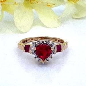 May include: A gold ring with a heart-shaped ruby surrounded by a halo of small diamonds. The ring also has two smaller ruby accents on either side of the center stone.