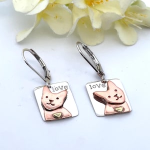 May include: A pair of silver earrings with a rectangular pendant featuring a pink cat design and the word "love". The cat has a gold heart on its chest. The earrings have a lever back closure.