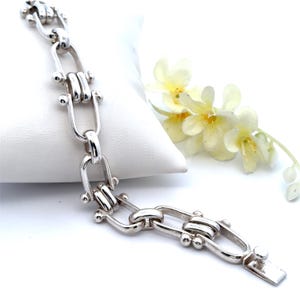 May include: A silver bracelet with a unique link design. The bracelet features a series of elongated, U-shaped links connected by smaller, double-bar links. Small, round accents are placed on the links. The bracelet is displayed on a white surface with yellow flowers in the background.