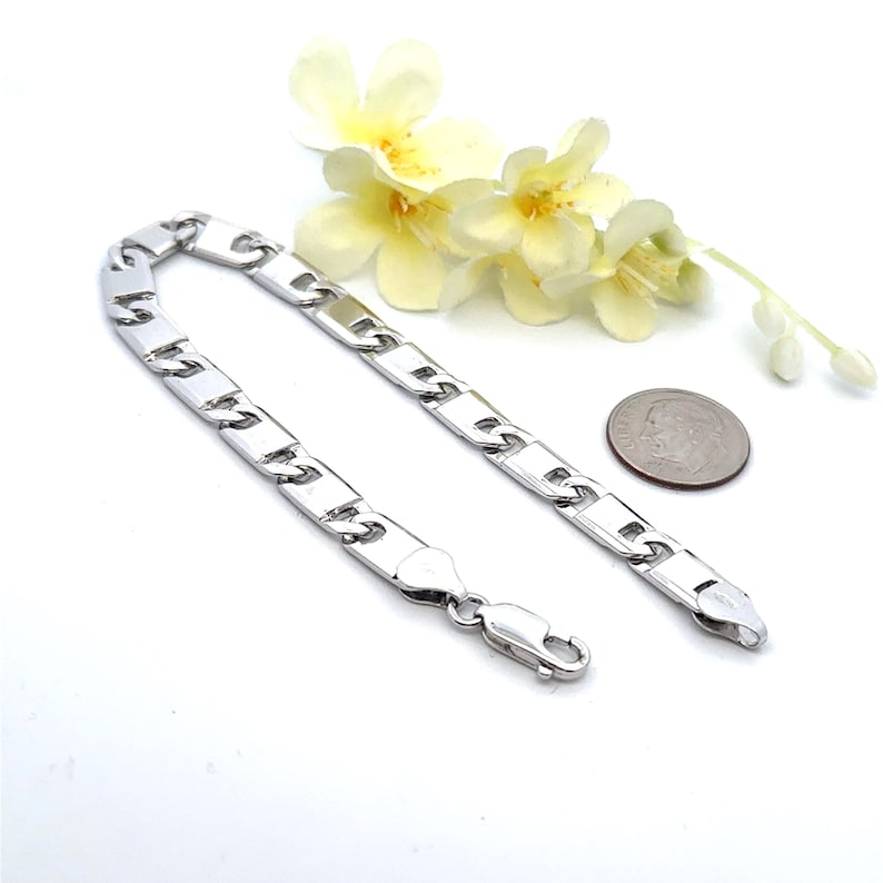 May include: A silver-colored bracelet with a rectangular link design. The bracelet is displayed on a white surface, next to a dime and a sprig of yellow flowers. The bracelet has a clasp closure.