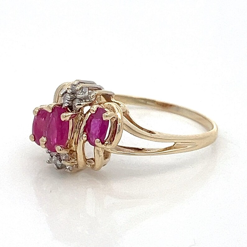May include: A gold ring featuring three oval-shaped ruby gemstones and smaller diamond accents. The ring has a delicate, open-work design.