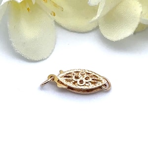 May include: A gold-colored filigree jewelry clasp, oval-shaped with a floral design in the center. The clasp has a small hook and loop for secure closure. The background is white with a blurred cream-colored flower.