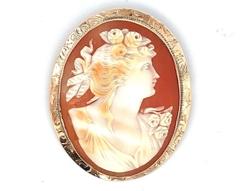 May include: A gold-toned cameo brooch featuring a woman's profile with a floral crown. The cameo is set in an oval frame with an intricate design.