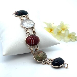 May include: A link bracelet featuring five oval-shaped stones carved with scarab beetle motifs. The stones include a dark black, a translucent clear, a banded reddish-brown, a mottled green, and a final dark black stone, all set in gold-toned metal.
