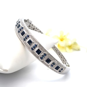 May include: A silver-toned hinged bangle bracelet with a geometric design. The bracelet features alternating square and oval dark blue gemstones, accented with smaller clear stones. The bracelet is displayed on a white surface with a yellow flower in the background.