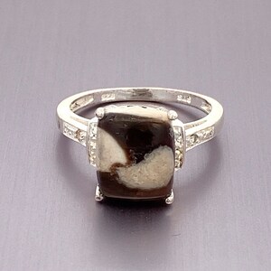 May include: A silver ring featuring a large, rectangular, brown and white stone set in the centre. The stone is surrounded by smaller, clear stones.