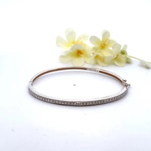 May include: A silver-colored bangle bracelet with small, clear stones set along the front. The bracelet is hinged and has a clasp. A few yellow flowers are in the background, set against a white background.