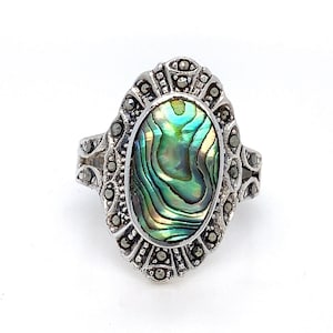 May include: A silver ring with an oval abalone shell set in a detailed, ornate design. The ring is decorated with small, black marcasite stones.