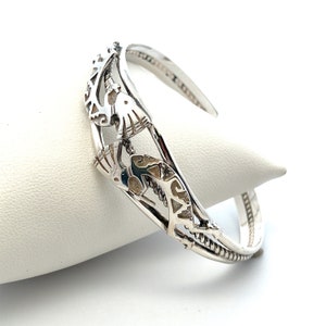 May include: Silver cuff bracelet with a detailed design of two birds in flight. The bracelet is made of polished silver and has a smooth, rounded finish.