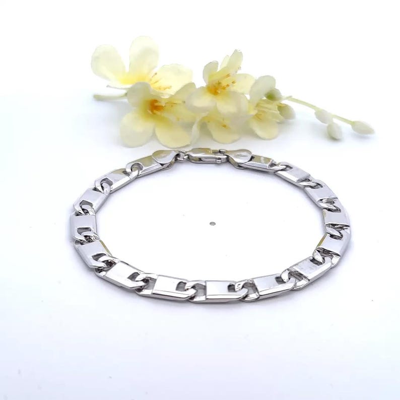 May include: A silver-colored bracelet with a rectangular link design. The bracelet is displayed on a white surface, with a sprig of yellow flowers in the background. The bracelet is a jewelry item.