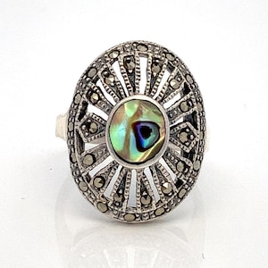 May include: A silver ring with a round, openwork design featuring a central abalone shell inlay. The ring is set with marcasite stones.