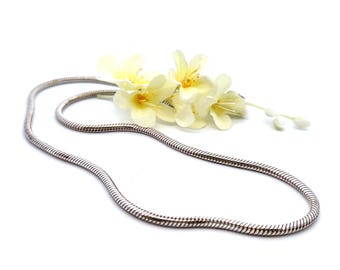 May include: A silver snake chain necklace on a white background with a cluster of white flowers.
