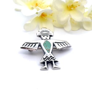 May include: A silver brooch shaped like a stylized bird figure with outstretched wings. The piece features a central teardrop-shaped inlay of green turquoise and black etched detailing on the wings, head, and skirt area of the figure.