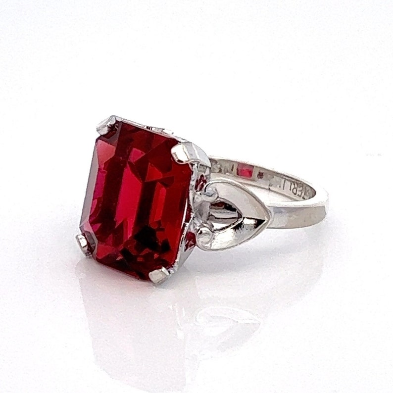 May include: A silver ring featuring a large, red, emerald-cut gemstone. The ring band is adorned with a heart-shaped design.