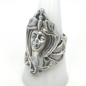 May include: A silver ring with a detailed design of a woman's face. The ring is made of a silver metal and has a detailed design of a woman's face. The ring is a statement piece and would be a great addition to any jewelry collection.