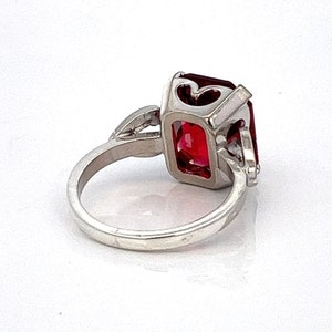 May include: A silver ring featuring a large, red, emerald-cut gemstone. The ring has a delicate, heart-shaped design on the band.