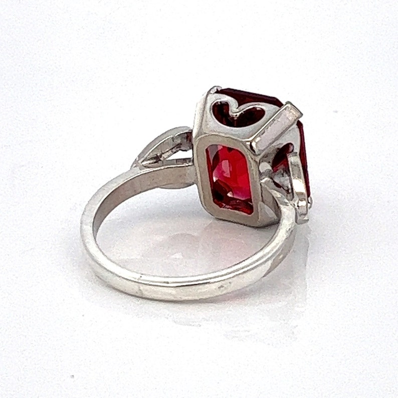 May include: A silver ring featuring a large, red, emerald-cut gemstone. The ring has a delicate, heart-shaped design on the band.