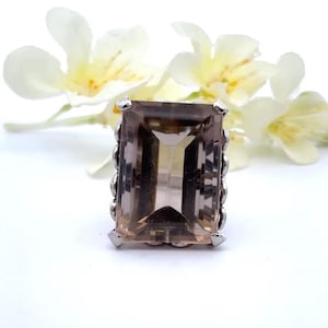 May include: A silver ring featuring a large, rectangular, smoky quartz gemstone. The gemstone has a light brown hue with visible internal facets. The ring's setting has a decorative, textured design around the stone, and is set against a white background with white flowers.