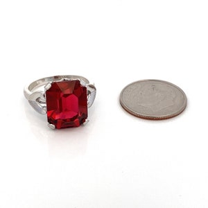 May include: A silver ring featuring a large, red, emerald-cut gemstone. The ring is set with two small, heart-shaped accents on either side of the main stone. The ring is shown next to a UK ten pence coin for size comparison.