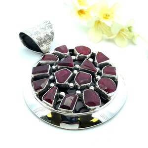 May include: A circular silver pendant featuring multiple irregular, deep red ruby gemstones set in a cluster. The pendant has a wide, polished silver border and a textured, patterned silver bail at the top for attaching to a chain or necklace.