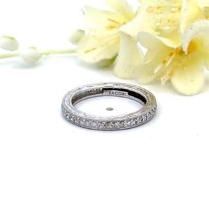 May include: A silver-toned metal eternity band featuring a continuous row of small, clear, round-cut stones set along the outer edge. The band has intricate engraved detailing on the sides and the word TACORI is stamped on the inner surface.