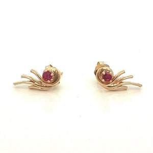 May include: A pair of gold earrings with a red gemstone in the center of each earring. The earrings have a delicate, flowing design.