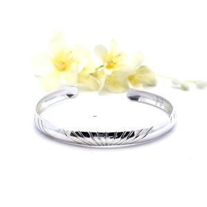 May include: A silver cuff bracelet with a textured, ridged design. The open-ended bracelet is displayed on a white surface, with yellow flowers in the background. The bracelet is a piece of jewelry.