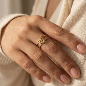 Puede incluir: Un anillo dorado con las palabras "fuck off" en escritura cursiva. El anillo se lleva en un dedo, y el diseño es visible sobre un fondo neutro. El anillo es una joya.