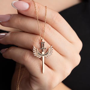 May include: A gold necklace with a pendant featuring a sword with wings. The sword and wings are adorned with black and white cubic zirconia stones.