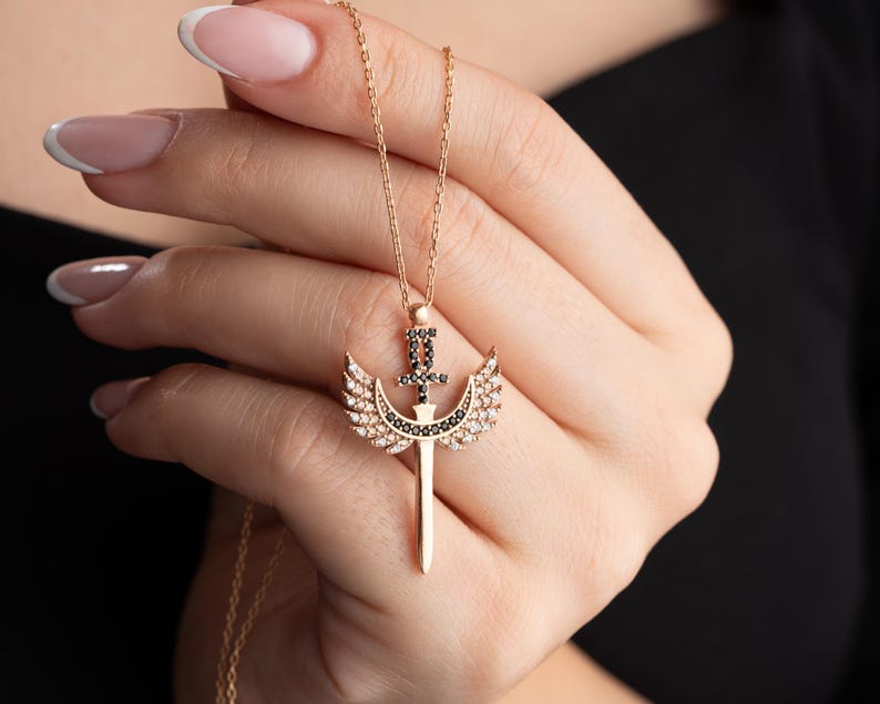 Puede incluir: Un collar de oro con un colgante que representa una espada con alas. La espada y las alas est&aacute;n adornadas con piedras de circonita c&uacute;bica negras y blancas.