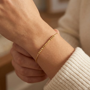 Puede incluir: Una delicada pulsera dorada con una serie de pequeñas cuentas redondas y barras rectangulares. La pulsera se lleva en la muñeca. El diseño es sencillo y elegante. Un accesorio elegante.