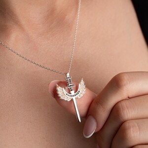 Puede incluir: Un collar de plata con un colgante que representa una espada con alas. La espada y las alas est&aacute;n adornadas con peque&ntilde;as piedras negras y blancas.